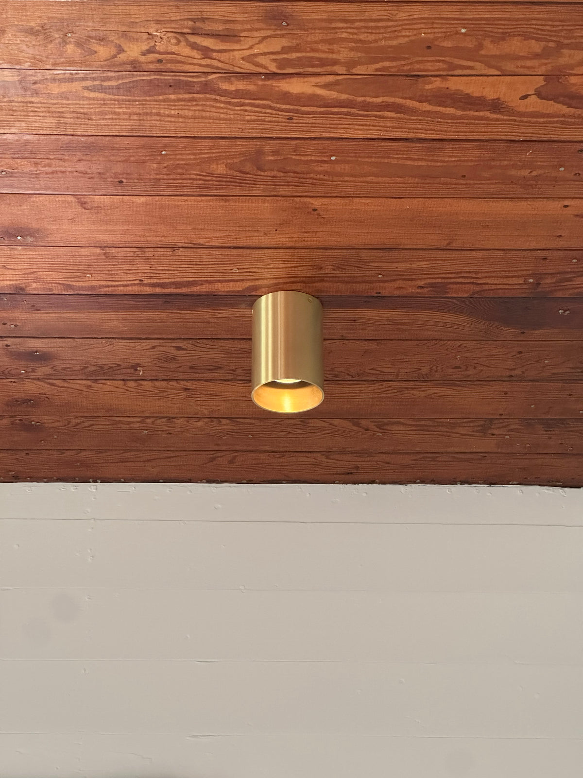The Beau Laiton Trimless Downlight by Long Made Co. features a minimalist gold brass cylinder, mounted on a wooden ceiling with the wall below painted white.