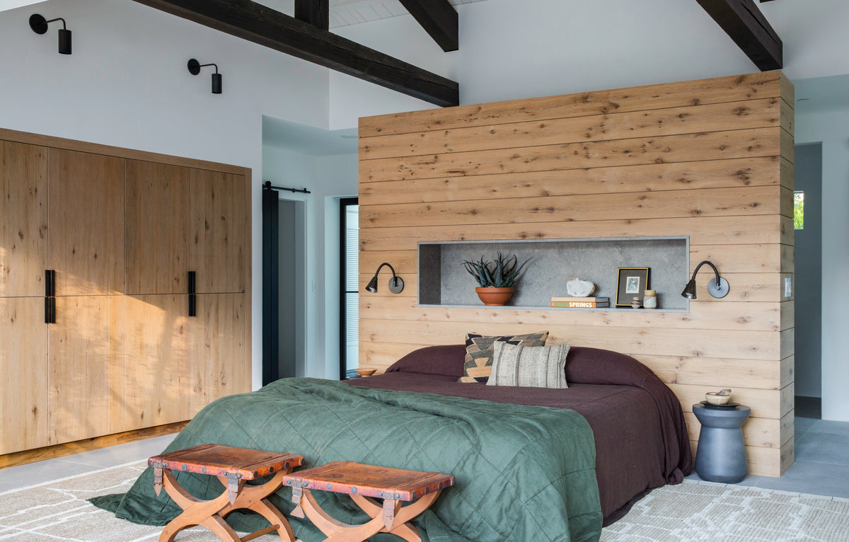 Modern bedroom with a wood-paneled accent wall behind a bed with green and brown bedding, two wooden benches at the foot, built-in shelf, large wardrobe on the left, and Cove Bent Wall Lamp by Long Made Co. highlighting exposed ceiling beams.