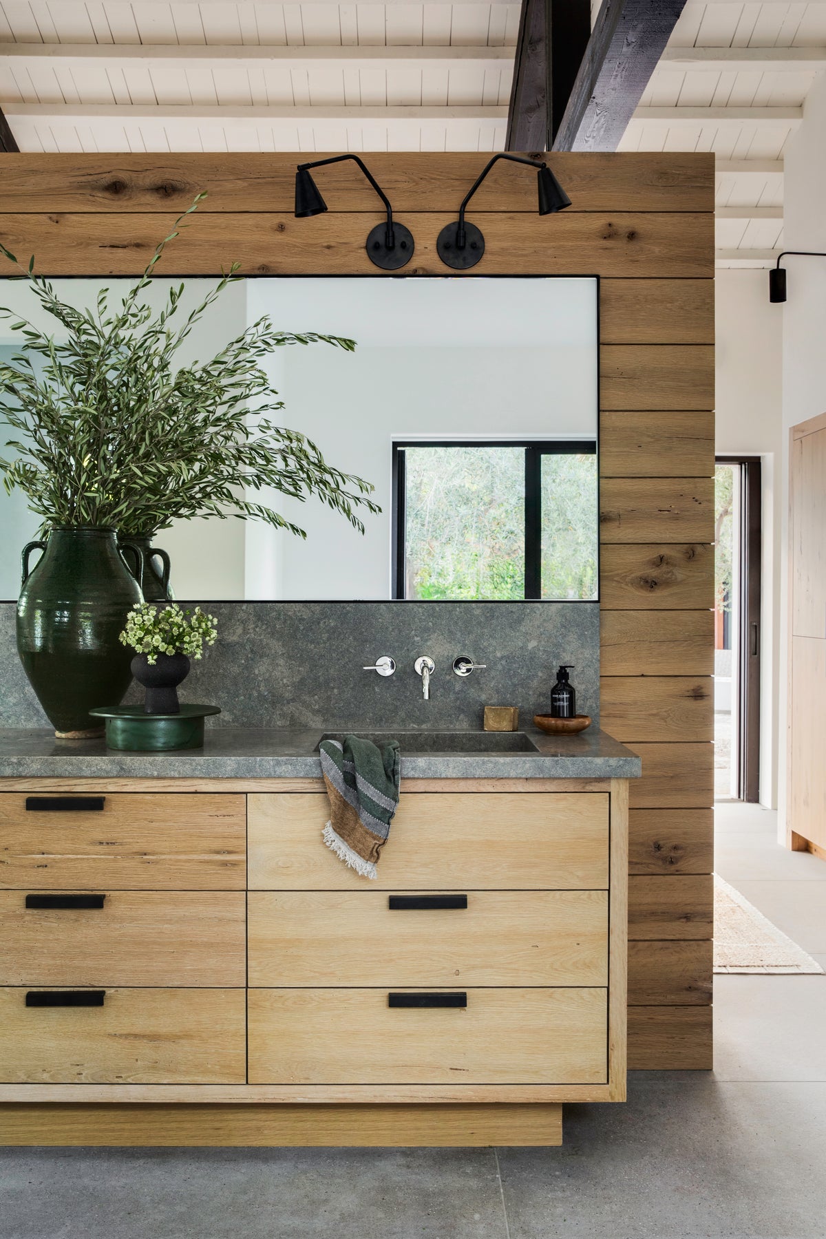 A modern bathroom with a wood accent wall, large mirror, concrete countertop, light wood drawers, green vase with tall branches, small plant, and the Tiel Arch Wall Lamp by Long Made Co. mounted above the mirror.
