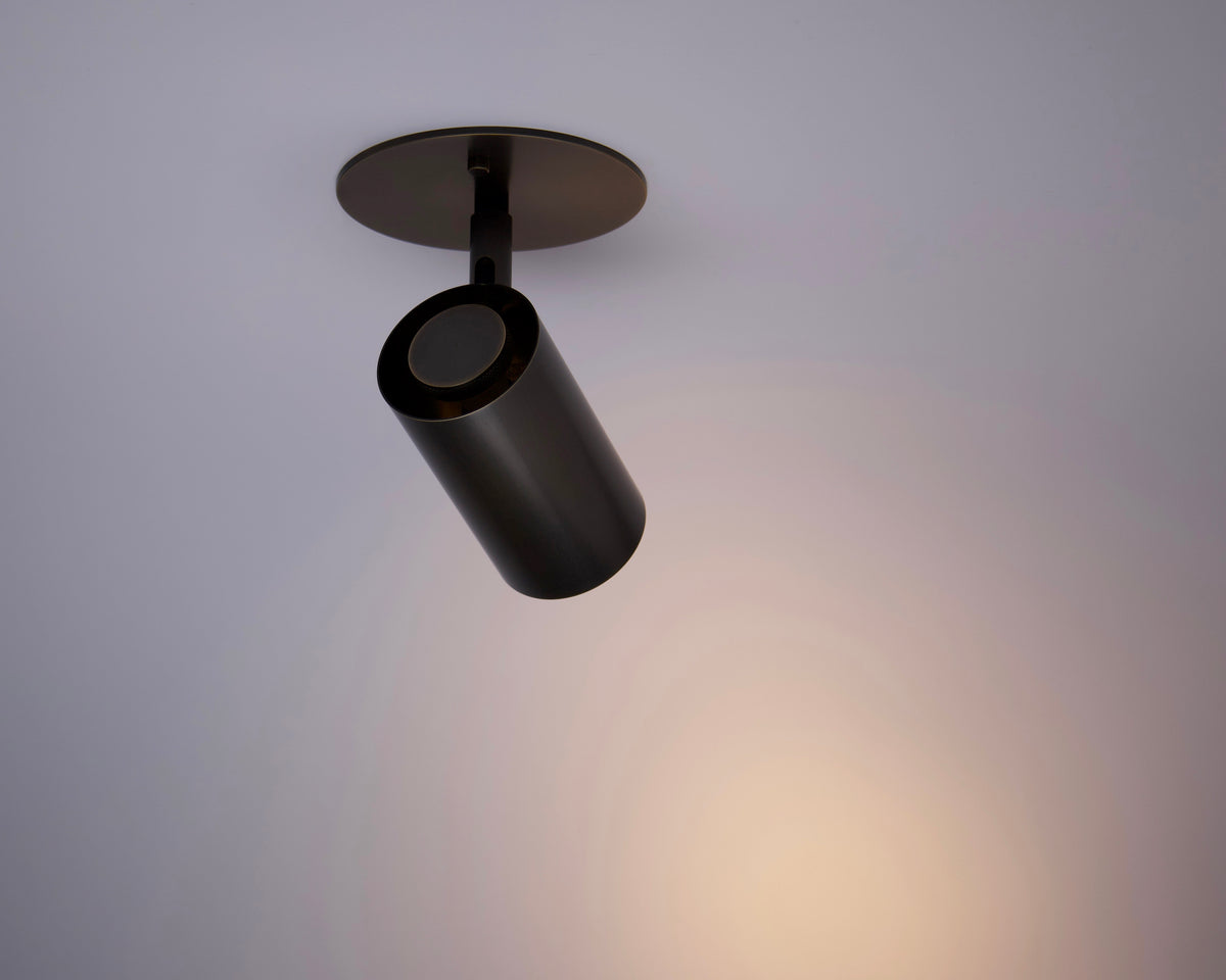The Brass Cylinder Adjustable Spot - Side Mount by Long Made Co. is turned on, casting a focused warm beam onto a pale ceiling. This modern, minimalistic fixture features an adjustable brass cylinder design for directional lighting.