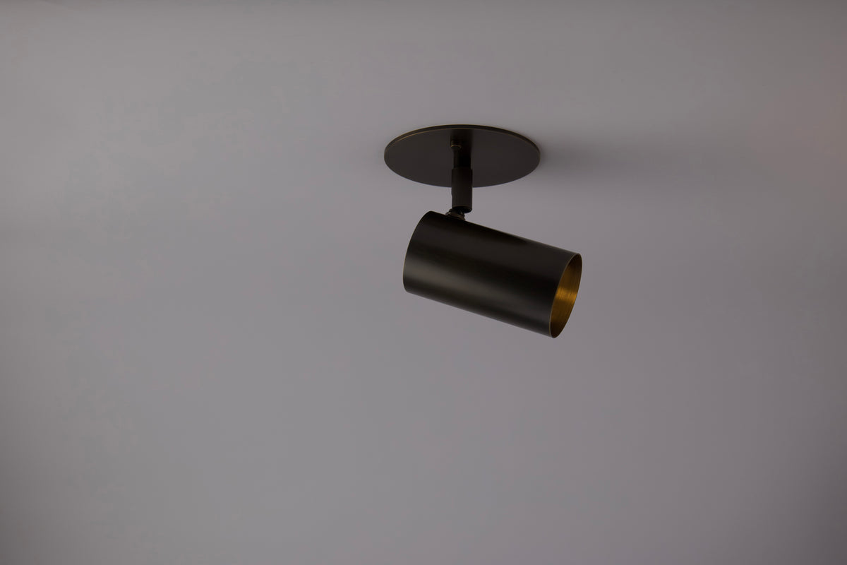 The Long Made Co. Brass Cylinder Adjustable Spot - Side Mount is mounted on a flat circular base against a light gray ceiling, angled downward and to the right, bringing sleek mid-century modern style to the space.
