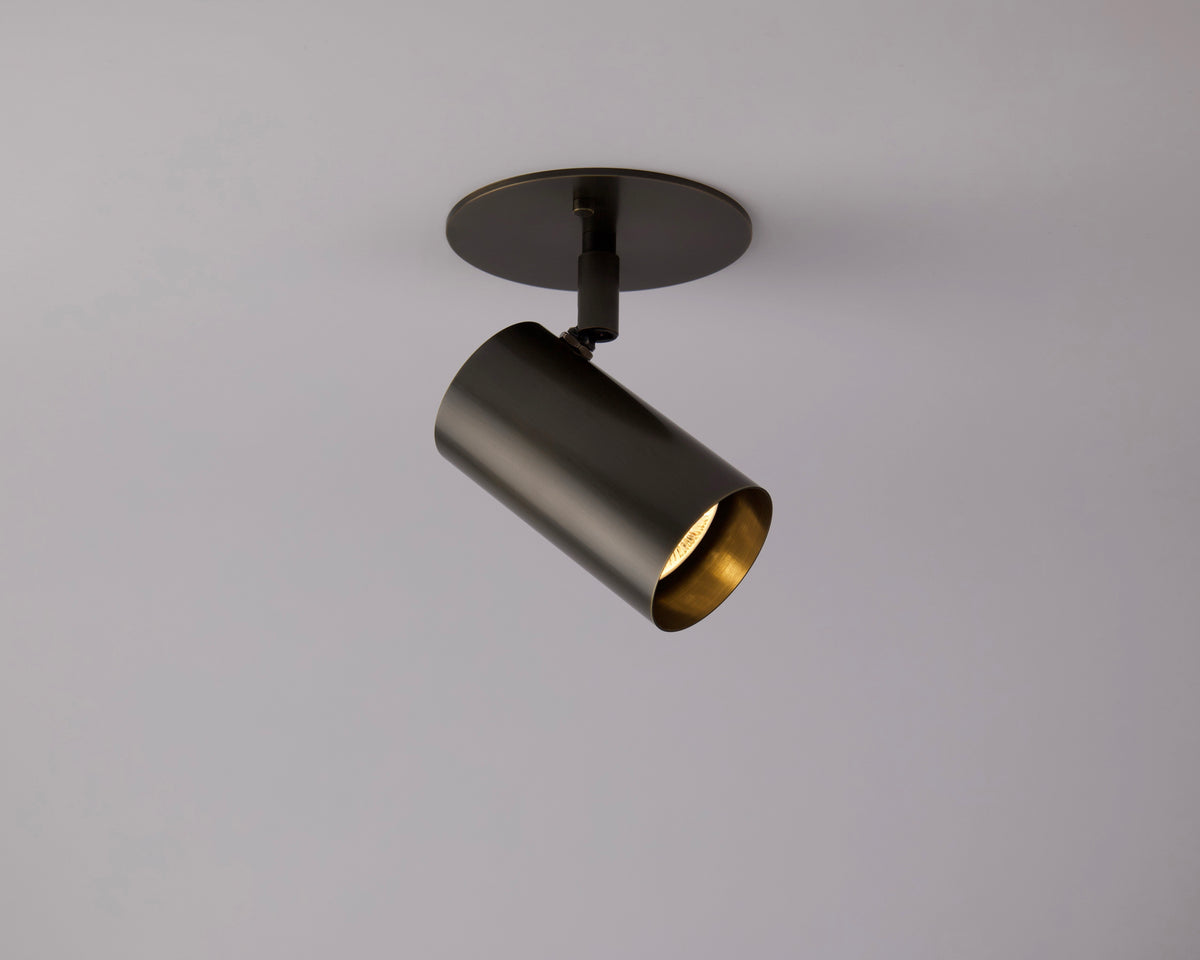 The Brass Cylinder Adjustable Spot - Side Mount by Long Made Co. is a modern black fixture with a cylindrical shape, mounted on a round base against a light gray background. Its spotlight angles downward, showcasing a gold-toned interior.