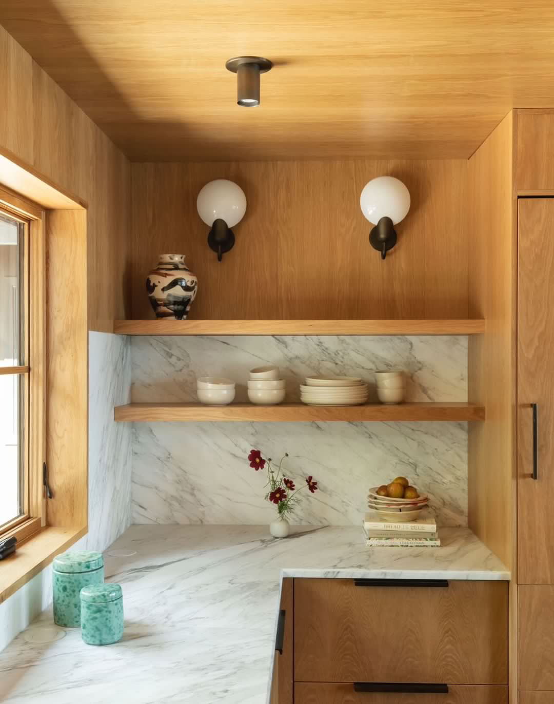A modern kitchen corner with wooden cabinets, marble backsplash and countertop, ceramic dishes, a vase of flowers, books, fruit, green glass jars, and the Brass Cylinder Flush Spot light fixture by Long Made Co.