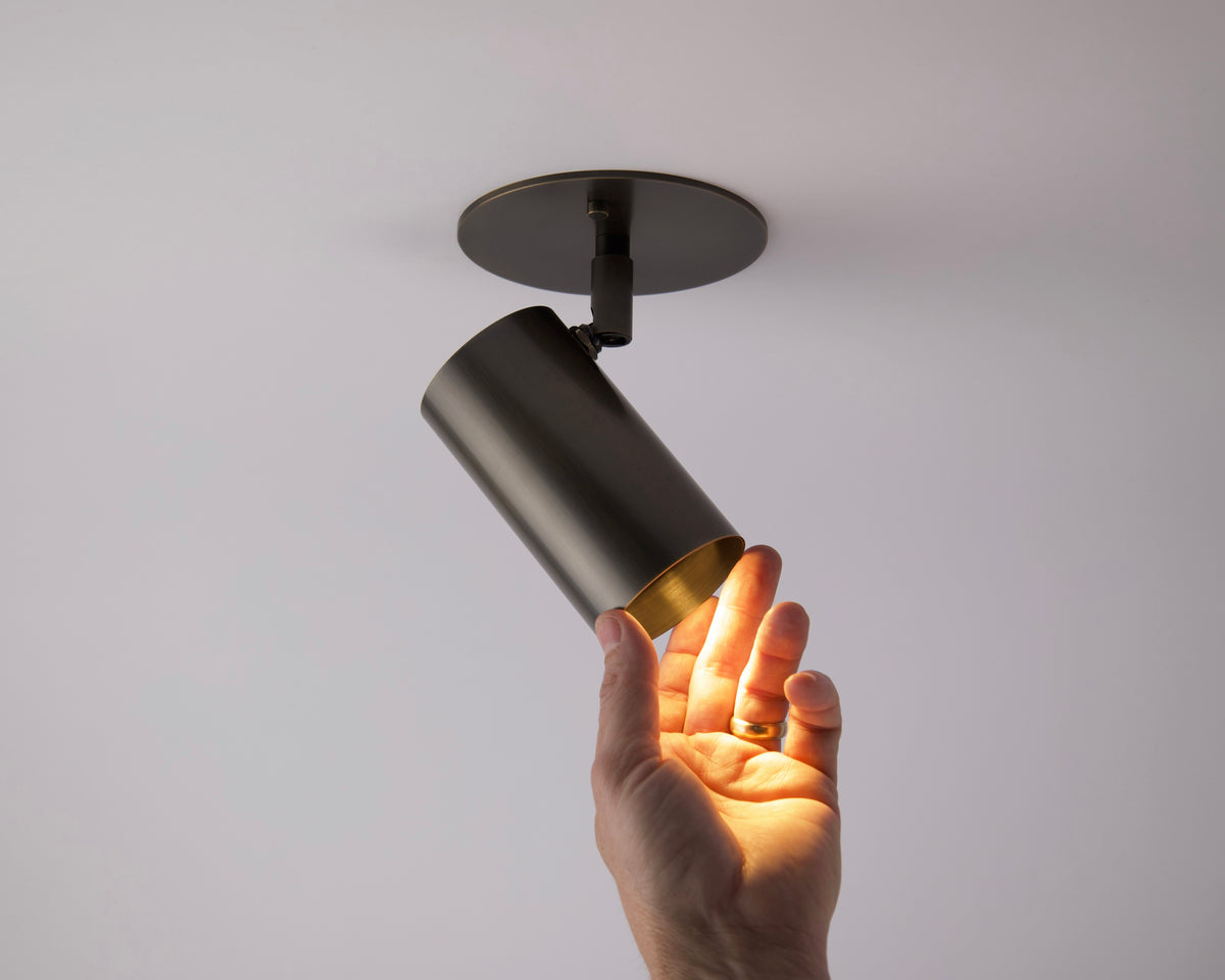 A hand reaches up to touch a Long Made Co. Brass Cylinder Adjustable Spot - Side Mount fixture, which is turned on and casts warm light against a plain white ceiling.