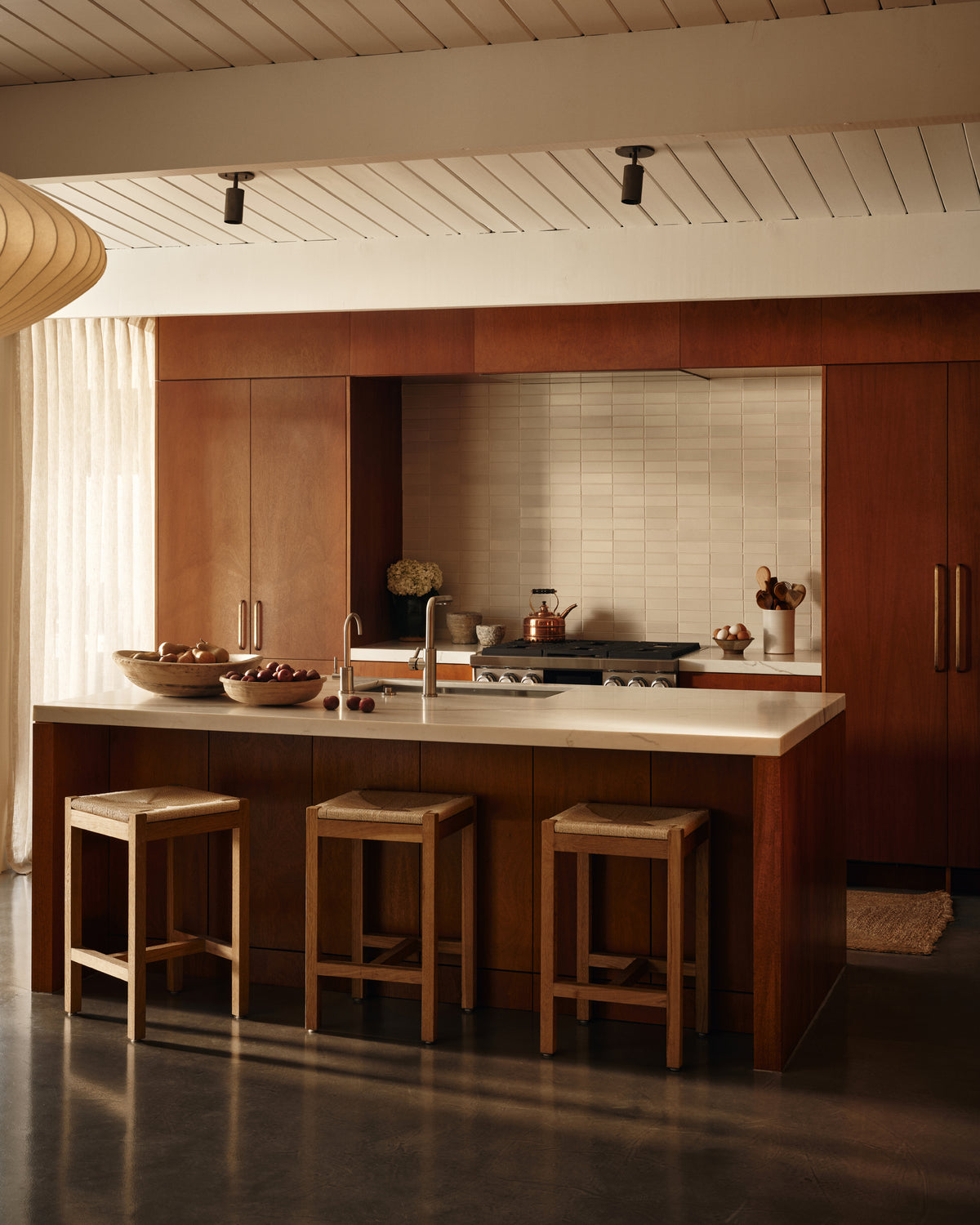 A modern kitchen features wooden cabinetry, a central island with a white countertop, three wooden stools, and Brass Cylinder Adjustable Spot - Top Mount lighting by Long Made Co., with natural light highlighting the stove and neutral-toned decor.
