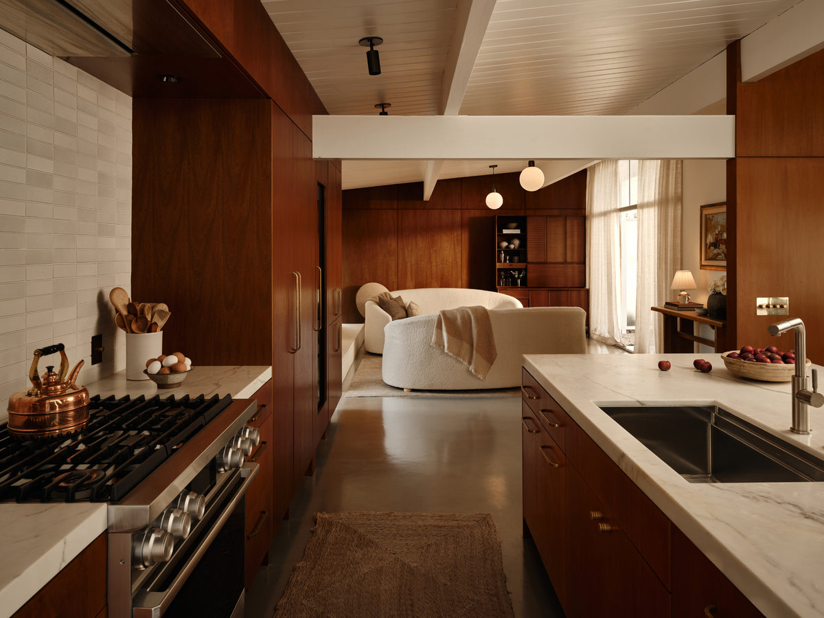 A modern kitchen flows into a cozy living room, featuring curved white sofas, wood cabinets, marble countertops, a gas stove, and is beautifully illuminated by the Brass Cylinder Adjustable Spot - Top Mount from Long Made Co. for a sophisticated touch.