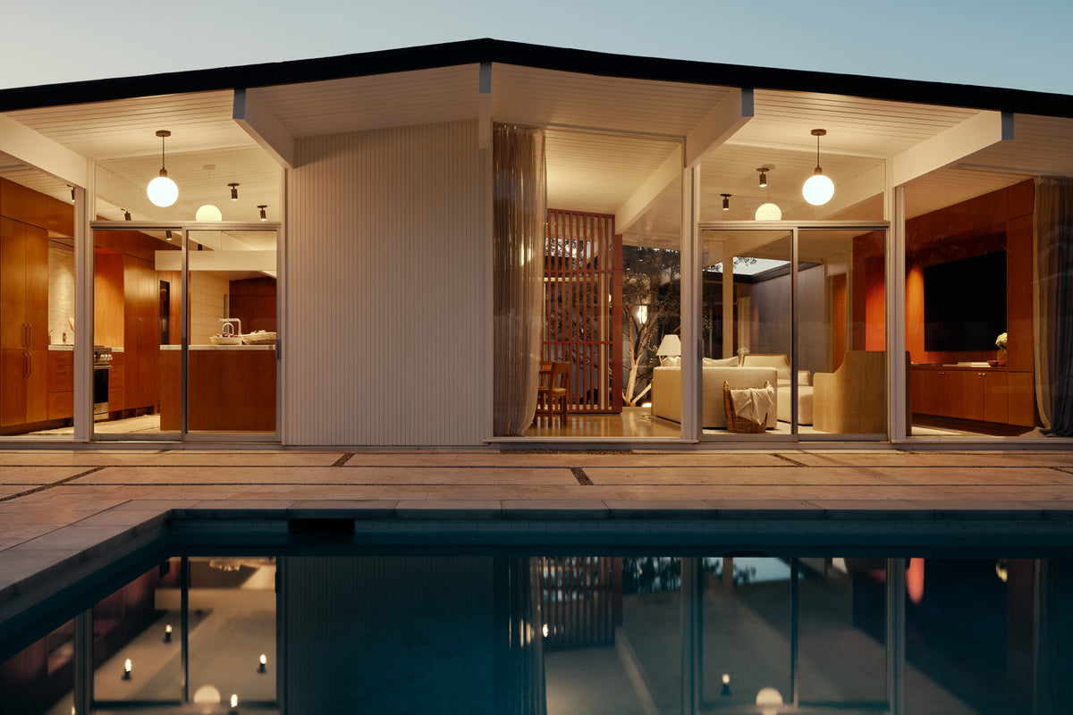 A modern house with large glass windows glows warmly at dusk from the Long Made Co. Brass Cylinder Adjustable Spot - Top Mount ceiling light, illuminating the open living area and kitchen. Outside, a swimming pool mirrors the home’s sleek architecture.