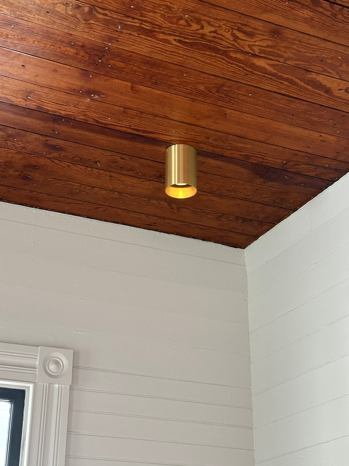 The Beau Laiton Trimless Downlight by Long Made Co., a gold cylindrical brass fixture, is mounted on a wooden ceiling above white paneled walls, with a window frame visible in the bottom left corner.