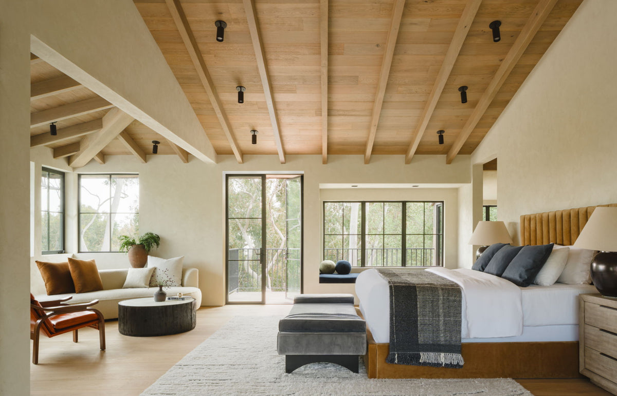 Spacious, modern bedroom with exposed wood ceiling beams, Long Made Co. Cove Adjustable Spot lighting above a large bed with gray and blue bedding, cozy window seating, light walls, and glass doors to a balcony overlooking greenery.