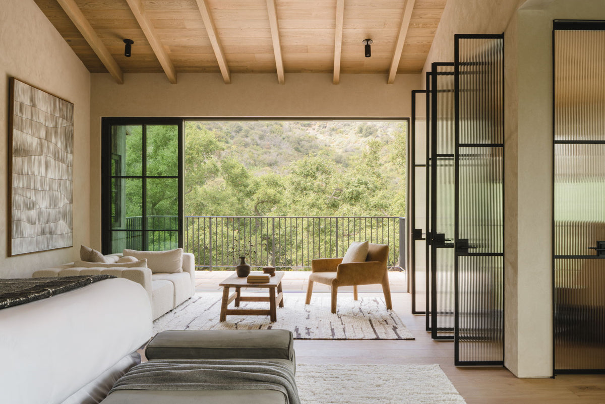 A modern living room with neutral tones, light wood ceiling, Cove Adjustable Spot lights by Long Made Co., large glass doors to a balcony with views of trees and hills, and sleek beige sofa, armchair, and coffee table.
