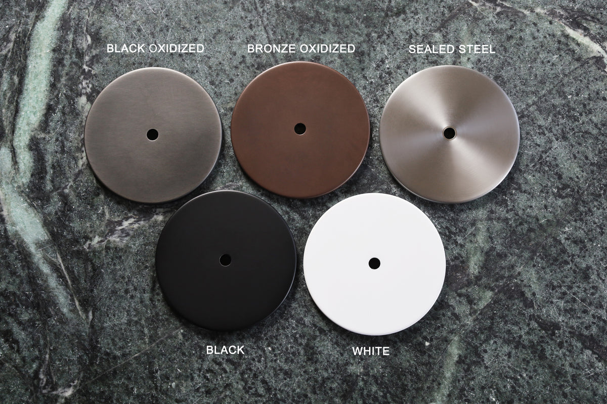 Five round metal discs for the Cove Cascade Drop Spot by Long Made Co. rest on green marble, labeled: Black Oxidized, Bronze Oxidized, Sealed Steel; bottom: Black, White—each with a central hole typical of adjustable ceiling spotlights.