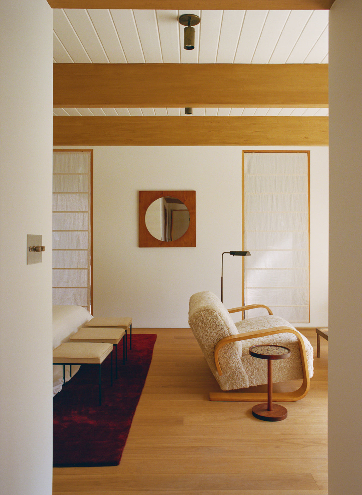 A modern living room with wooden beams, a cream sofa, a round side table, benches with cushions, a red rug, and a wooden-framed wall mirror between tall translucent panels is enhanced by Long Made Co.'s Brass Cylinder Adjustable Spot light.