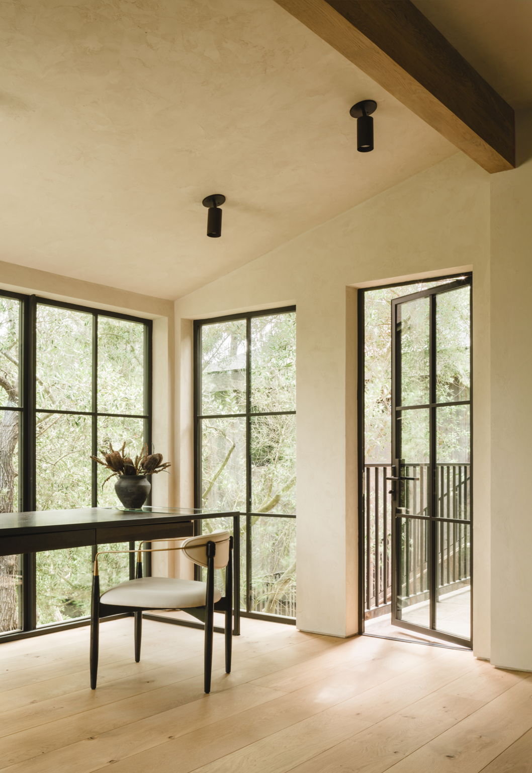 Minimalist office with light wood floors, black desk and chair, large black-framed windows, potted plant on the desk, and Cove Adjustable Spot by Long Made Co. for lighting. A door opens to a small balcony overlooking greenery.