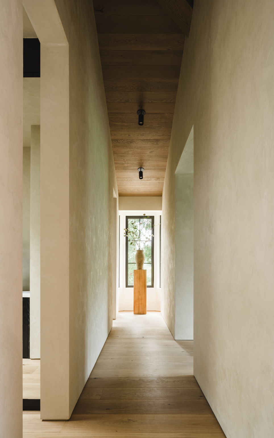 Minimalist hallway with cream walls and light wood floors, illuminated by Long Made Co.'s Highland Adjustable Spot lights. Soft natural light fills the space, ending at a window with a potted plant on a wooden pedestal.