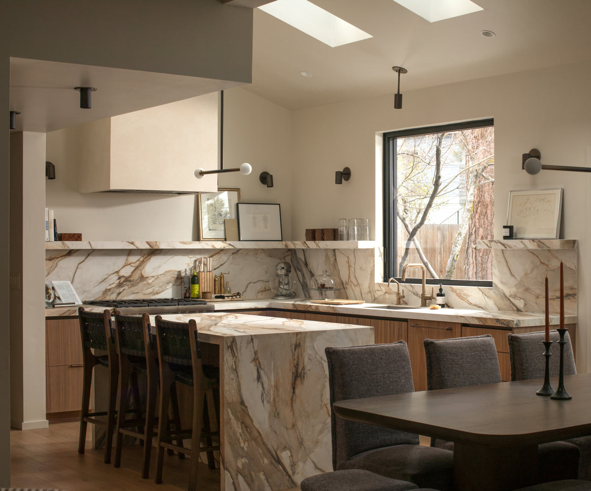 A modern kitchen features marble counters, wood cabinets, bar stools at an island, a dining table, large window, and skylights—finished with Long Made Co.'s Brass Cylinder Adjustable Spot - Side Mount for a mid-century modern touch.