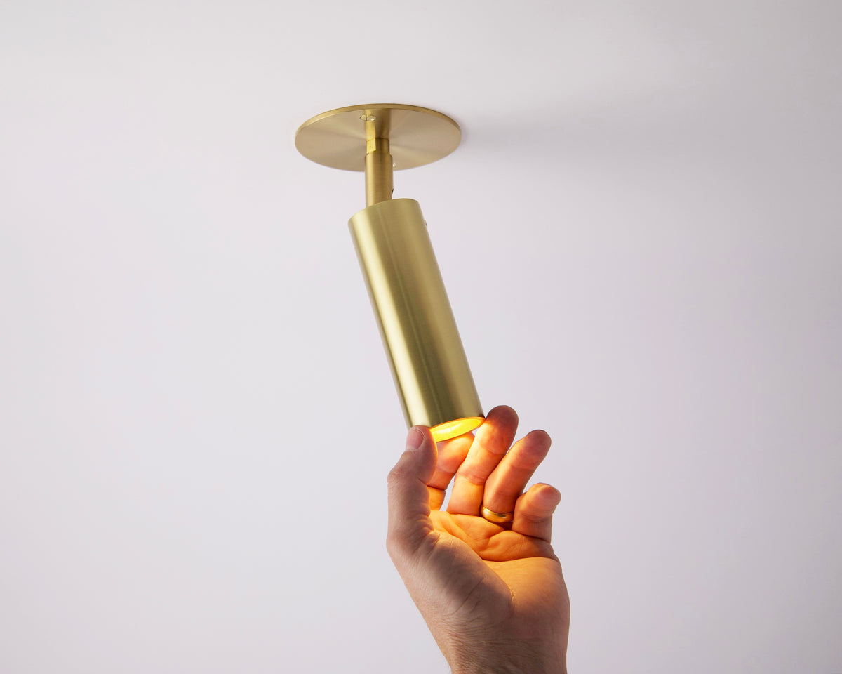 A hand reaches up to touch the Long Made Co. Micro Adjustable Spot with Long Shade in unlacquered brass, switched on and casting a warm glow against a plain white ceiling, showcasing its minimalist, modern design.