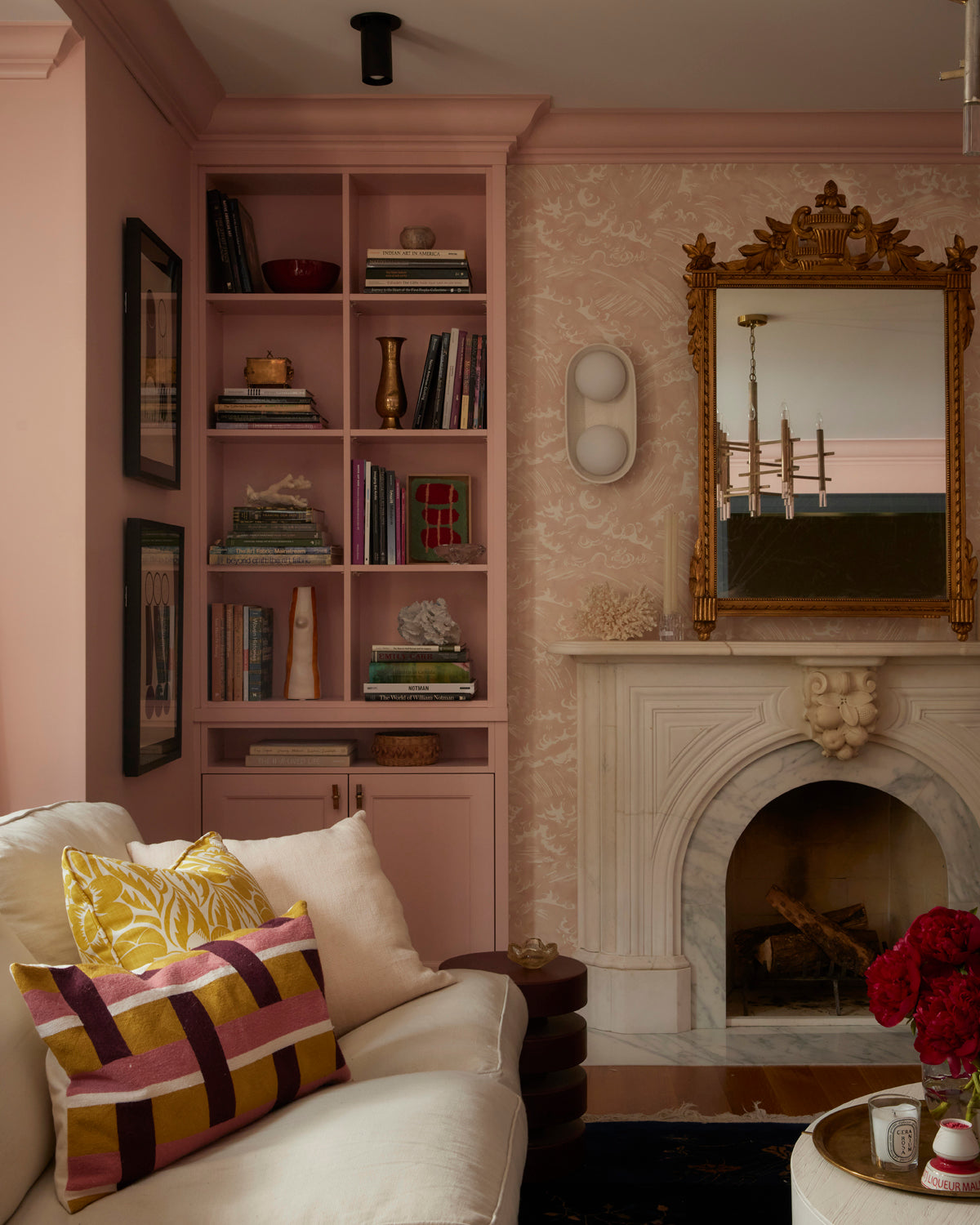 A cozy living room with blush pink walls, a white marble fireplace, a gold mirror, pink bookshelves, and a cream sofa with colorful pillows. Highland Flush Spot lights by Long Made Co. add warmth above red flowers on the table in front.