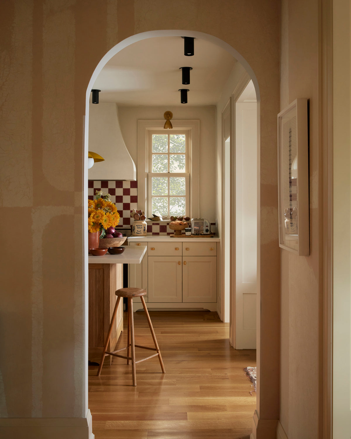 A cozy, sunlit kitchen with wooden floors, cream cabinets, a red and white checkered backsplash, sunflowers in a vase, and Long Made Co.'s Highland Flush Spot brass lights above a wooden stool under an arched doorway.