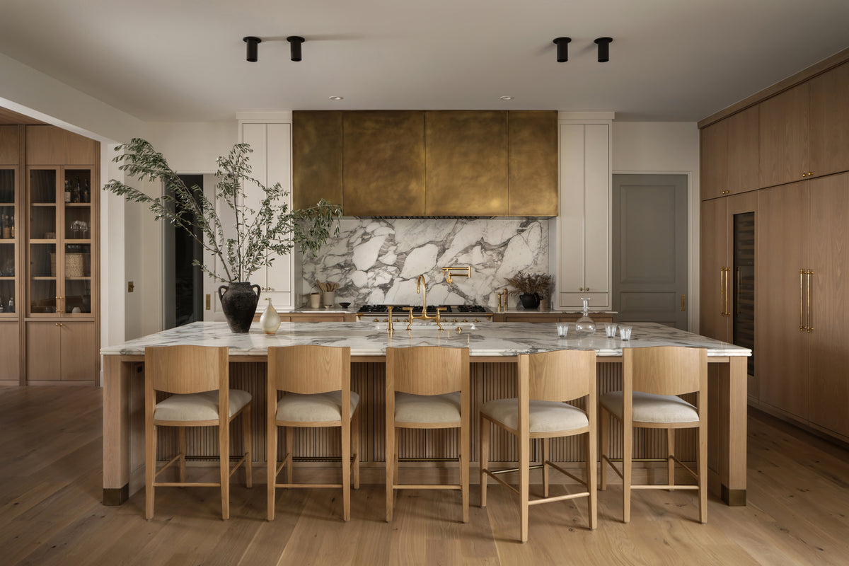 A modern kitchen with light wood cabinets, a large marble island with four wooden chairs, brass accents, and Long Made Co.'s Highland Flush Spot lighting illuminating a tall vase with branches on the marble island.
