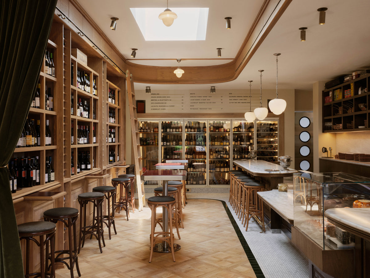 A cozy wine bar with shelves of wine bottles, high stools, wooden tables, and a display counter, featuring warm lighting from Long Made Co.’s Beau Laiton Monopoint minimalist brass fixtures and a welcoming atmosphere.
