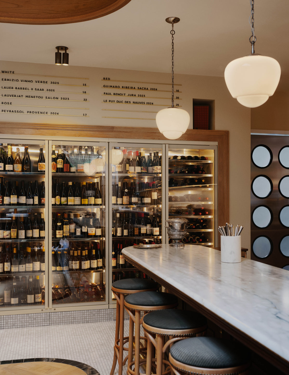 A stylish bar interior with a marble countertop, wooden bar stools, and a large glass wine fridge filled with bottles. Menu items are displayed on the wall above, illuminated by sleek mid-century modern lighting and an adjustable Brass Cylinder Double / Triple Spot by Long Made Co. Round pendant lights and solid brass ceiling downlights add a warm, inviting glow from the ceiling.