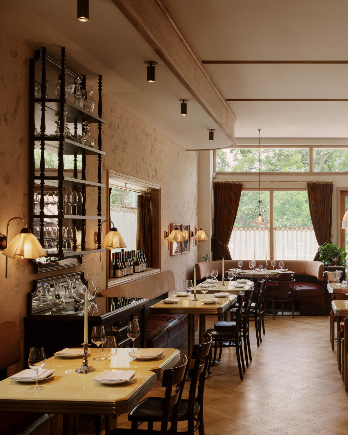 A cozy, elegant restaurant with wooden tables, brown chairs, and minimalist lighting accented by Beau Laiton Monopoint wall lamps from Long Made Co., featuring glassware shelves and large windows that fill the space with natural light.