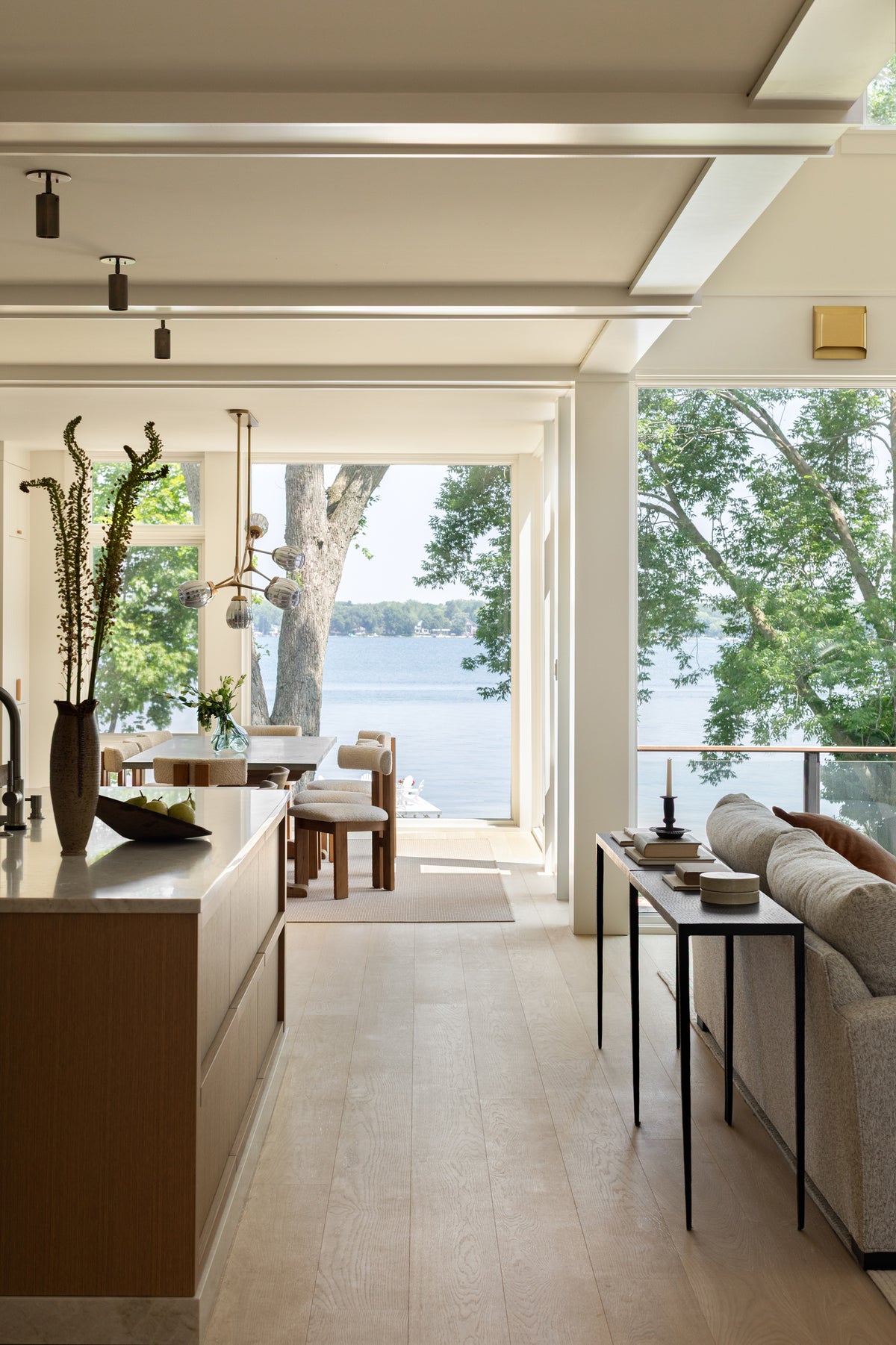 Modern open-plan space with light wood floors, neutral furniture, and large windows framing a scenic lake view. The Brass Cylinder Adjustable Spot - Top Mount by Long Made Co. adds mid-century flair and enhances the bright, airy room with dining table and island.