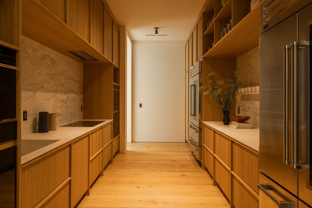 A modern kitchen features wooden cabinets, marble counters and backsplash, built-in appliances, open shelves, a floral vase, and cozy warm lighting from the Long Made Co. Brass Cylinder Adjustable Spot - Top Mount. A door is visible at the room’s end.