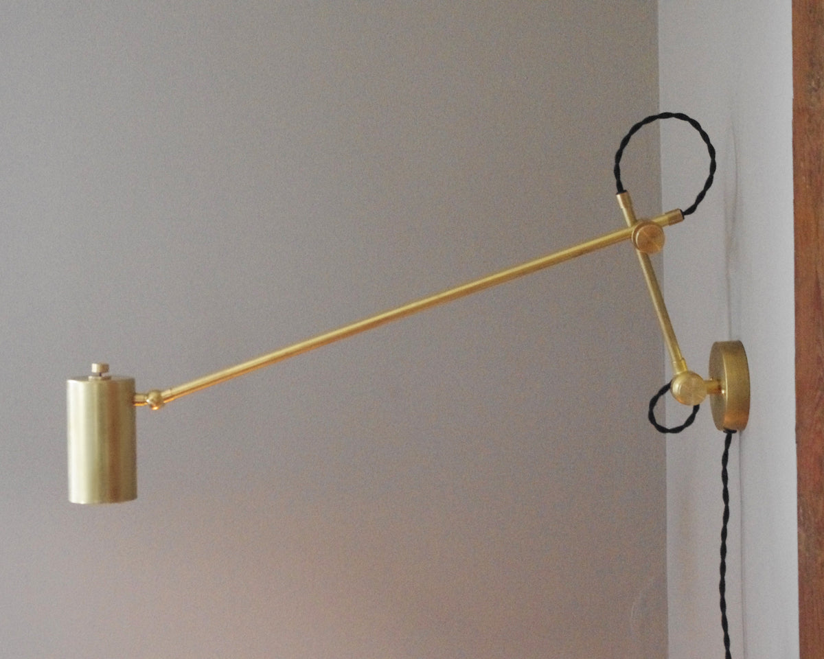 The Task Articulating Wall Lamp XL by Long Made Co. features a modern minimalist design with an adjustable brass body, cylindrical shade, and black twisted cord—ideal for stylish task lighting on a white wall near wood accents.