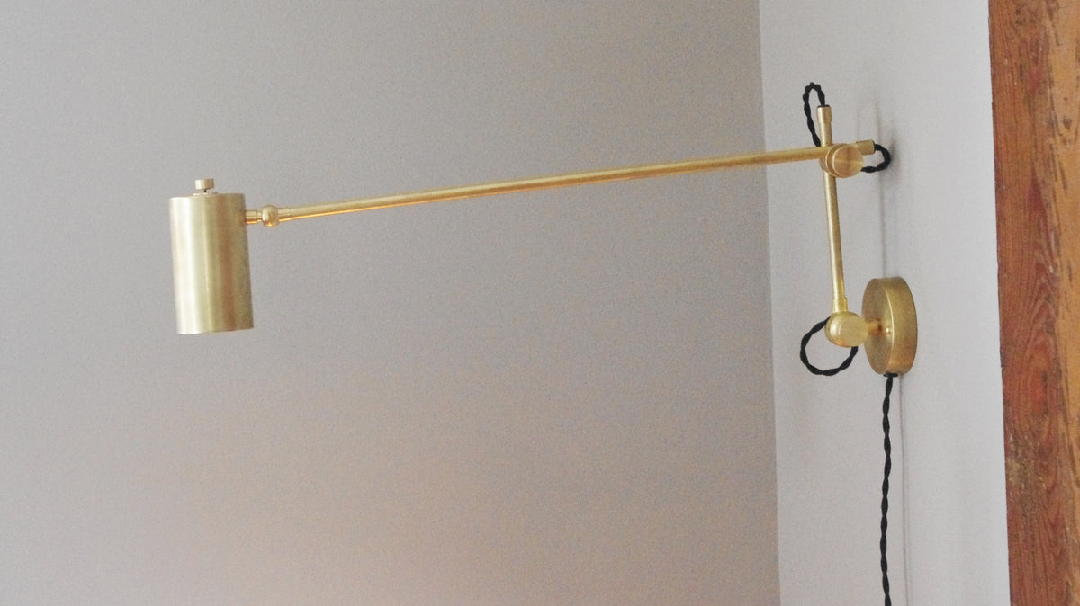 The Task Articulating Wall Lamp XL by Long Made Co. features a gold brass finish, cylindrical shade, and black twisted cord on a white wall with visible wood trim—ideal for stylish task lighting.