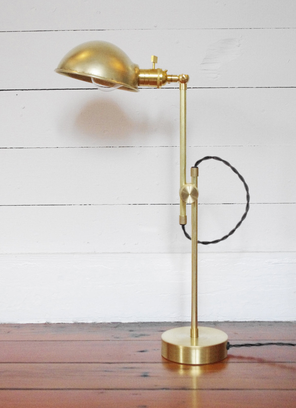 The Task Articulating Table Lamp by Long Made Co. features a brass finish, dome-shaped shade, and articulating arm. It sits on a wooden floor against a white planked wall, with its twisted black cord visible along the stand.
