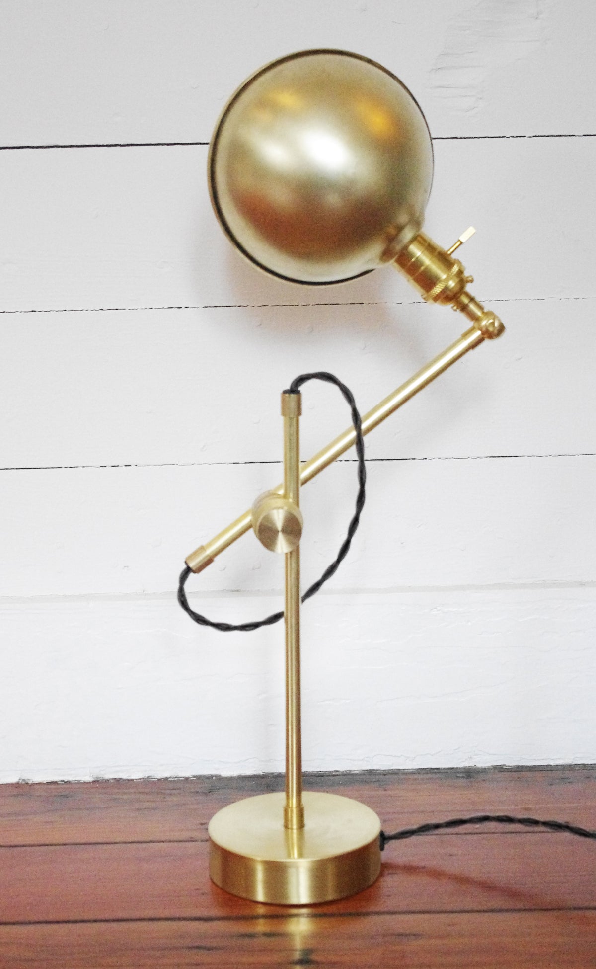 The Task Articulating Table Lamp by Long Made Co. features a modern brass design with a round adjustable head and black twisted cord, displayed on a wooden surface against a white paneled wall.