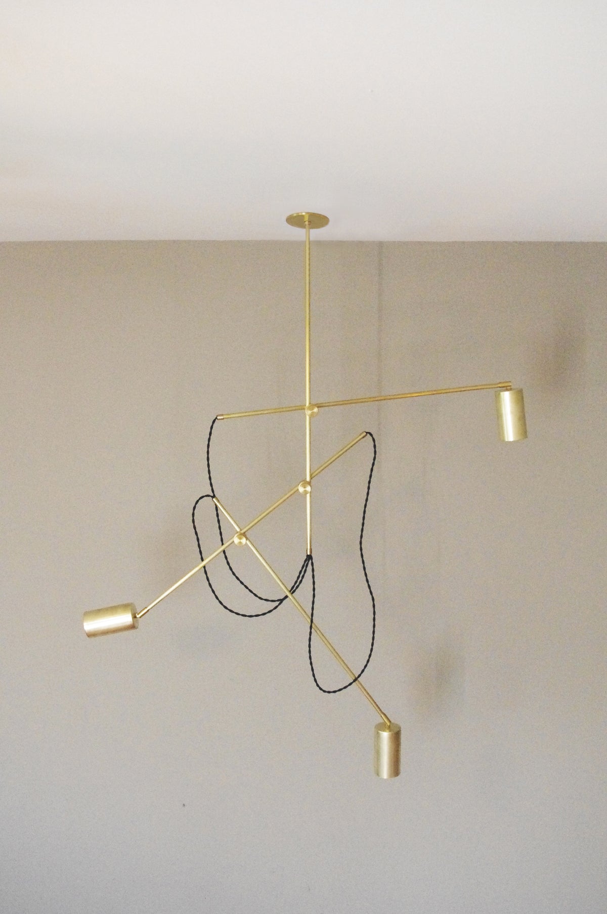 The Task Articulating Chandelier by Long Made Co. features three cylindrical brass lights on adjustable arms, suspended from a white ceiling. A black cord wraps around the fixture, set against a simple beige wall.