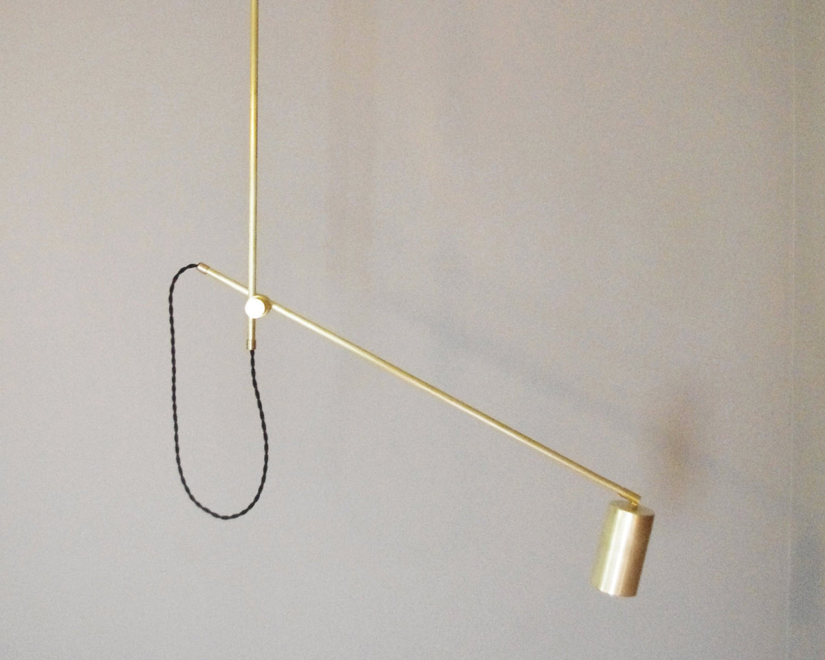 The Task Articulating Chandelier by Long Made Co. features a minimalist brass design with a slim, angular arm and cylindrical spotlight, suspended from the ceiling by a black cord for modern elegance in any space.