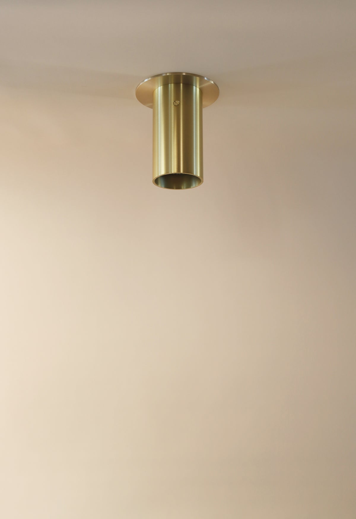 The Beau Laiton Flush Spot by Long Made Co. is a minimalist solid brass spotlight with a cylindrical design, mounted on a smooth beige ceiling, casting a subtle circular shadow around its base.
