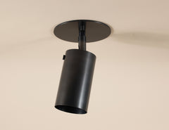 The Long Made Co. Brass Cylinder Adjustable Spot - Top Mount, with a round base, is mounted on a beige ceiling and angled downward for focused illumination.