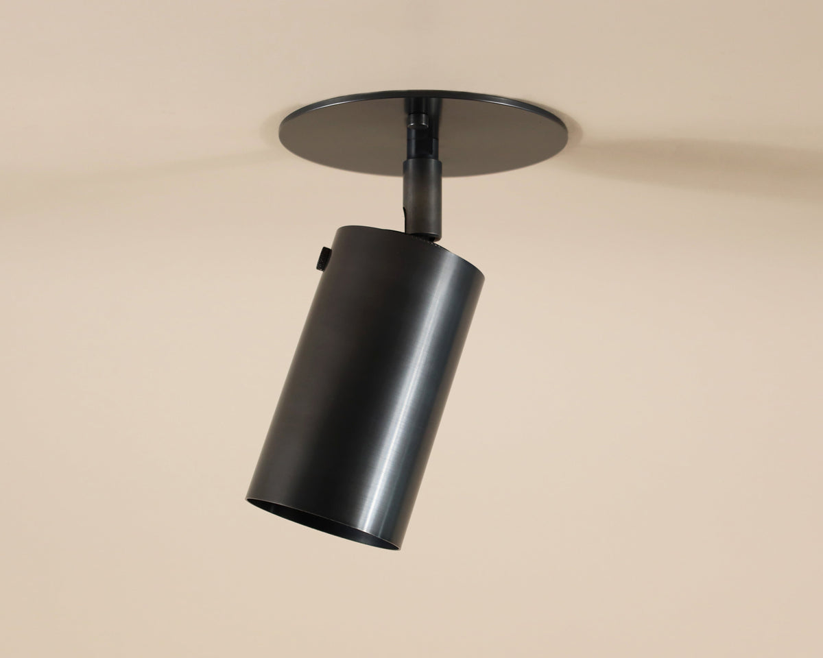The Brass Cylinder Adjustable Spot - Top Mount by Long Made Co. is a cylindrical directional downlight with an adjustable head, mounted on a round base against a beige ceiling.