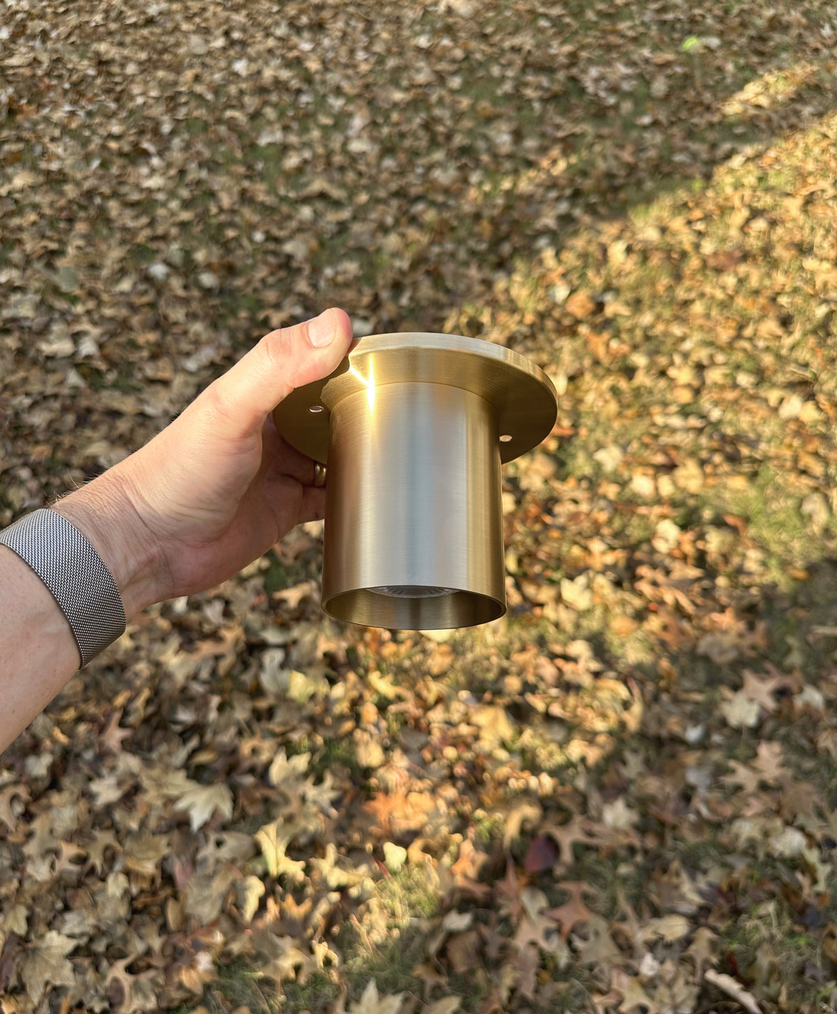 A hand holds the Long Made Co. Highland Flush Spot - Stocky, a solid brass cylinder light with a flange, outdoors above dry autumn leaves.