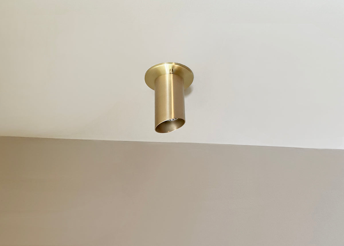 A Long Made Co. Highland Angle Spot, finished in gold and crafted from heavy-gauge solid brass, is installed as a flush monopoint on a white ceiling above a beige wall.