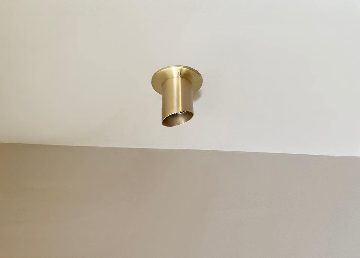 The Highland Angle Spot - Stocky by Long Made Co. is a modern solid brass flush ceiling fixture with a minimalist cylinder shape, mounted on a white ceiling above a beige wall for general and task lighting.