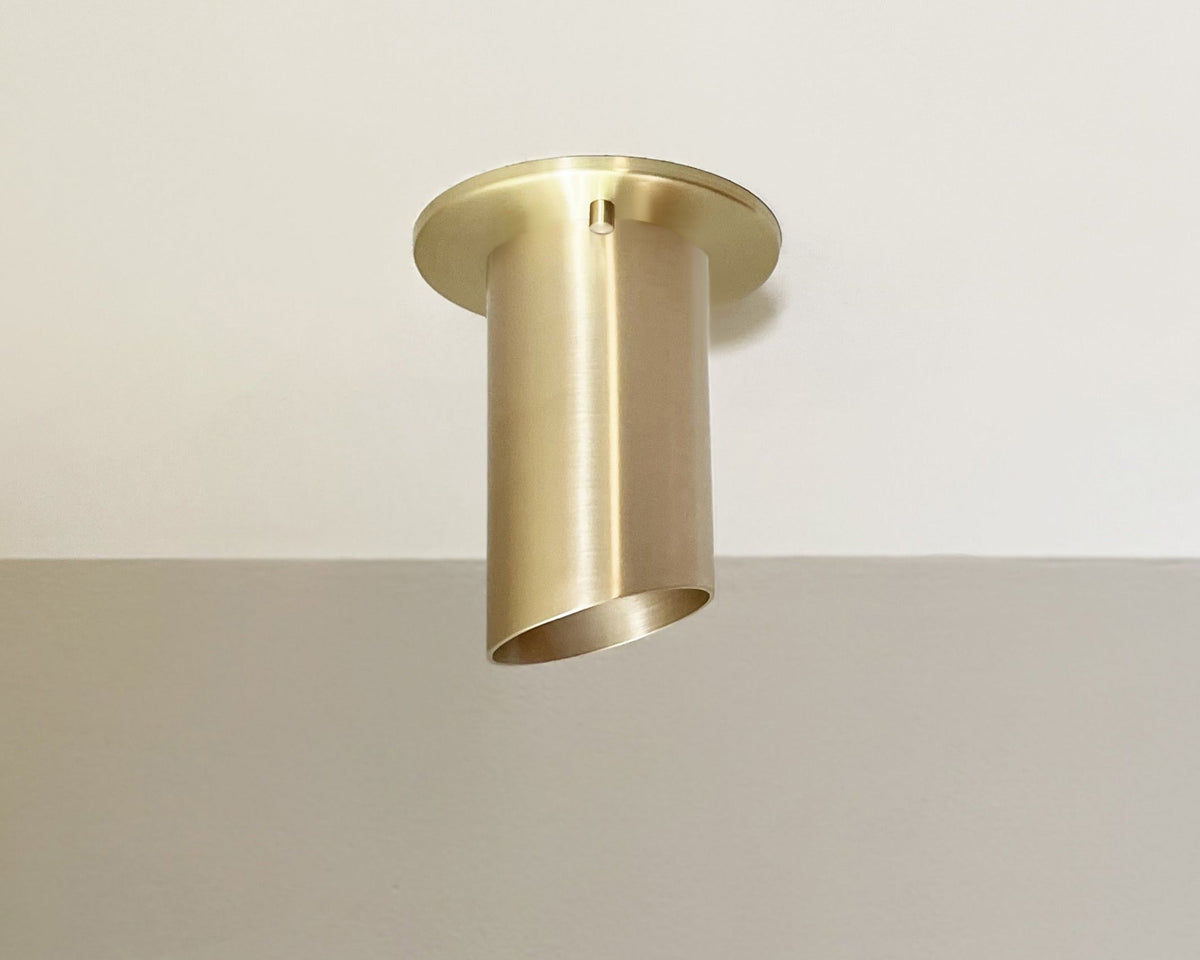 The Highland Angle Spot by Long Made Co., a modern cylinder light in heavy gauge solid brass, is flush-mounted on a white ceiling above a pale beige wall.