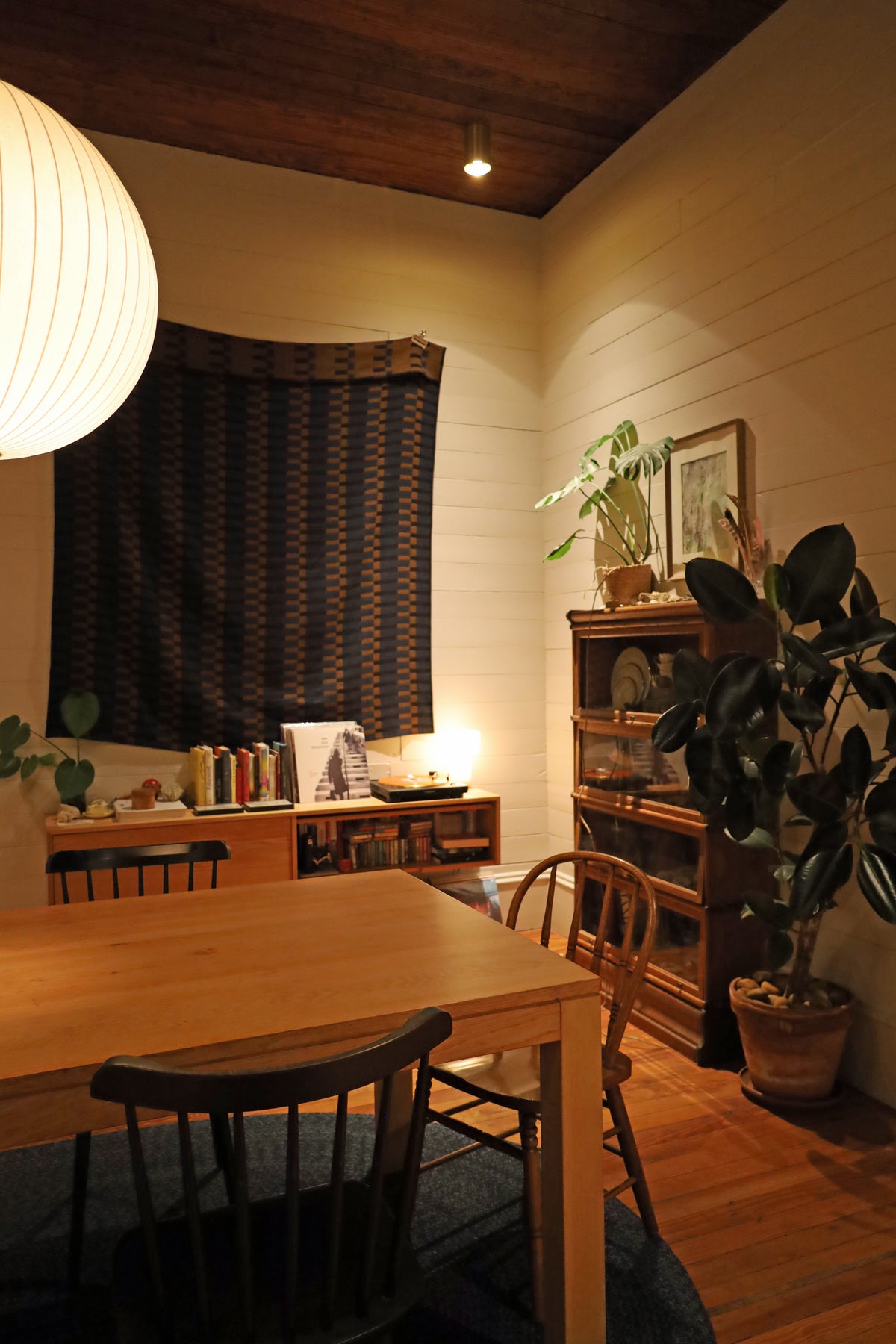 A cozy room with warm lighting and a wooden dining table, featuring a tall bookshelf with plants, books, and art. The Beau Laiton Trimless Downlight by Long Made Co. adds subtle elegance to the patterned wall hanging and cream walls.