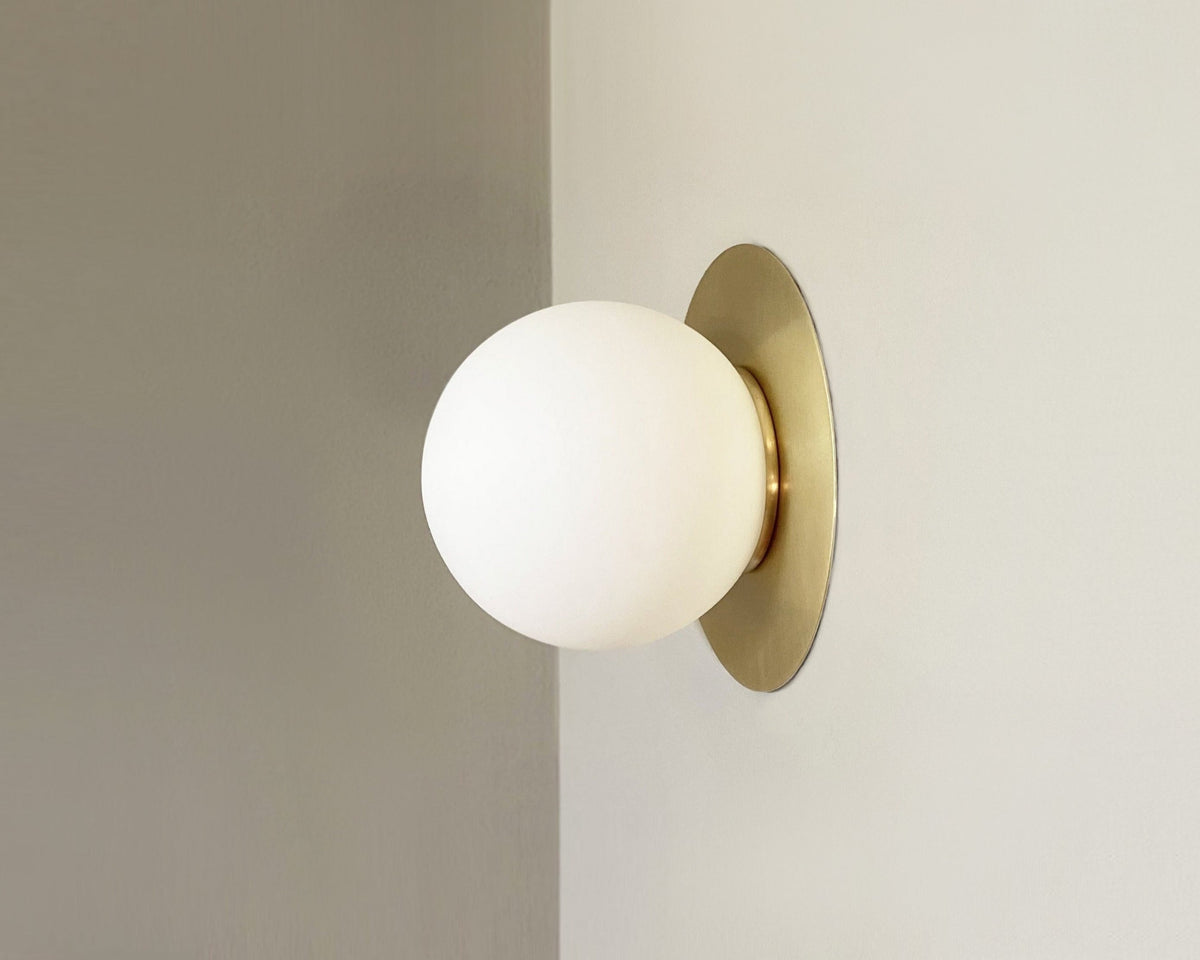 A wall-mounted light fixture with a gold-colored backplate and a white spherical opal shade, viewed from a side angle against a neutral background.
