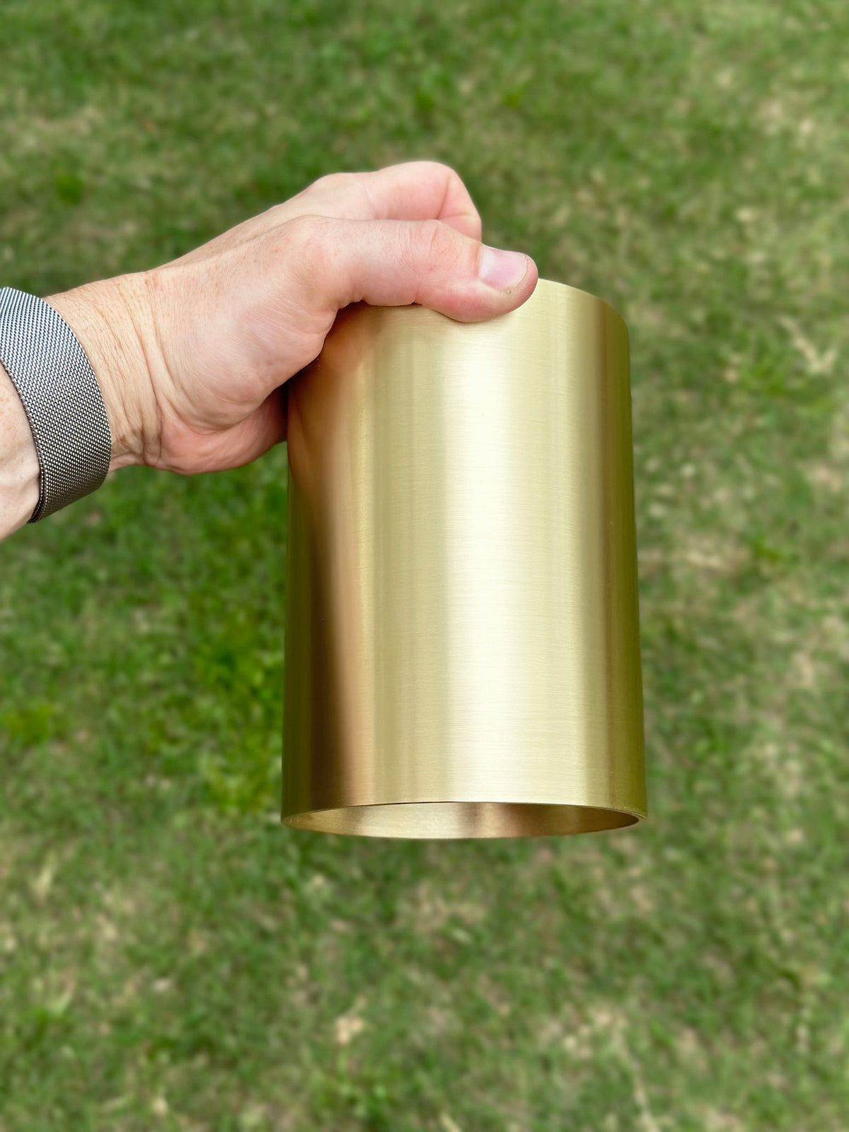 A hand holds the Long Made Co. Beau Laiton Trimless Downlight—a minimalist, cylindrical brass fixture—against a grassy outdoor backdrop.