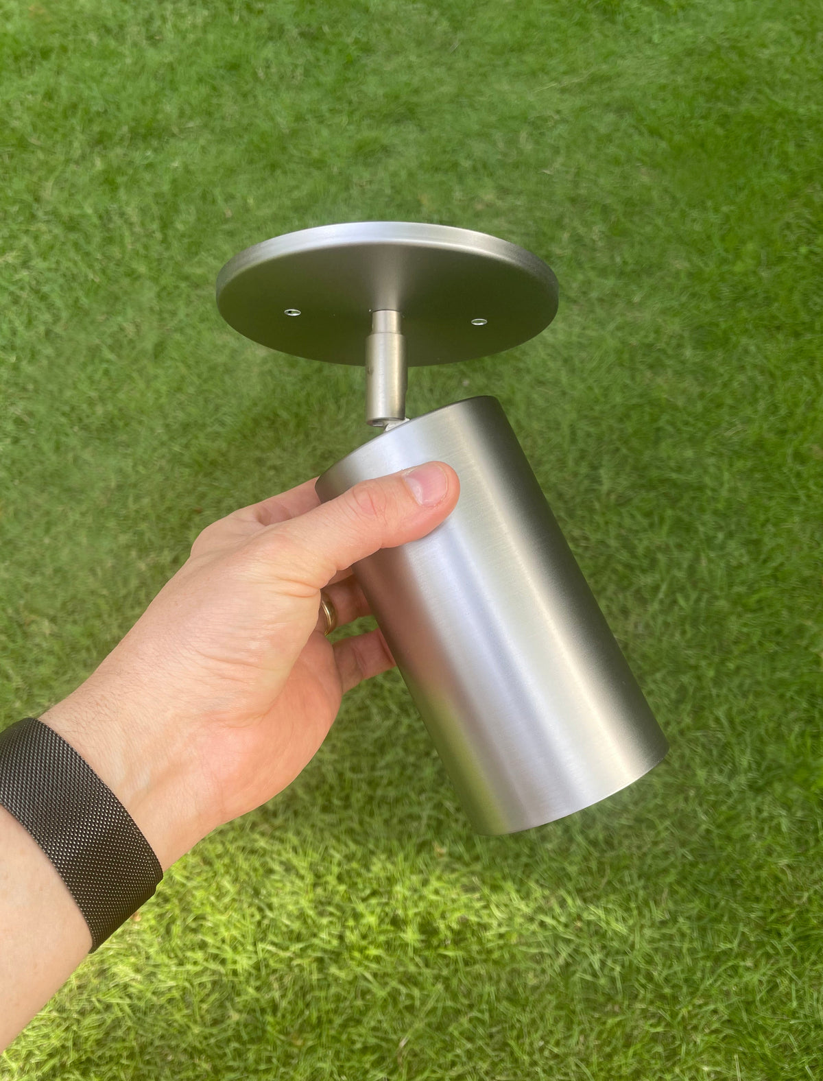 A hand holds the Cove Adjustable Spot by Long Made Co., a solid steel downlight with a round mounting plate, ideal for minimalist spotlights, against green grass.