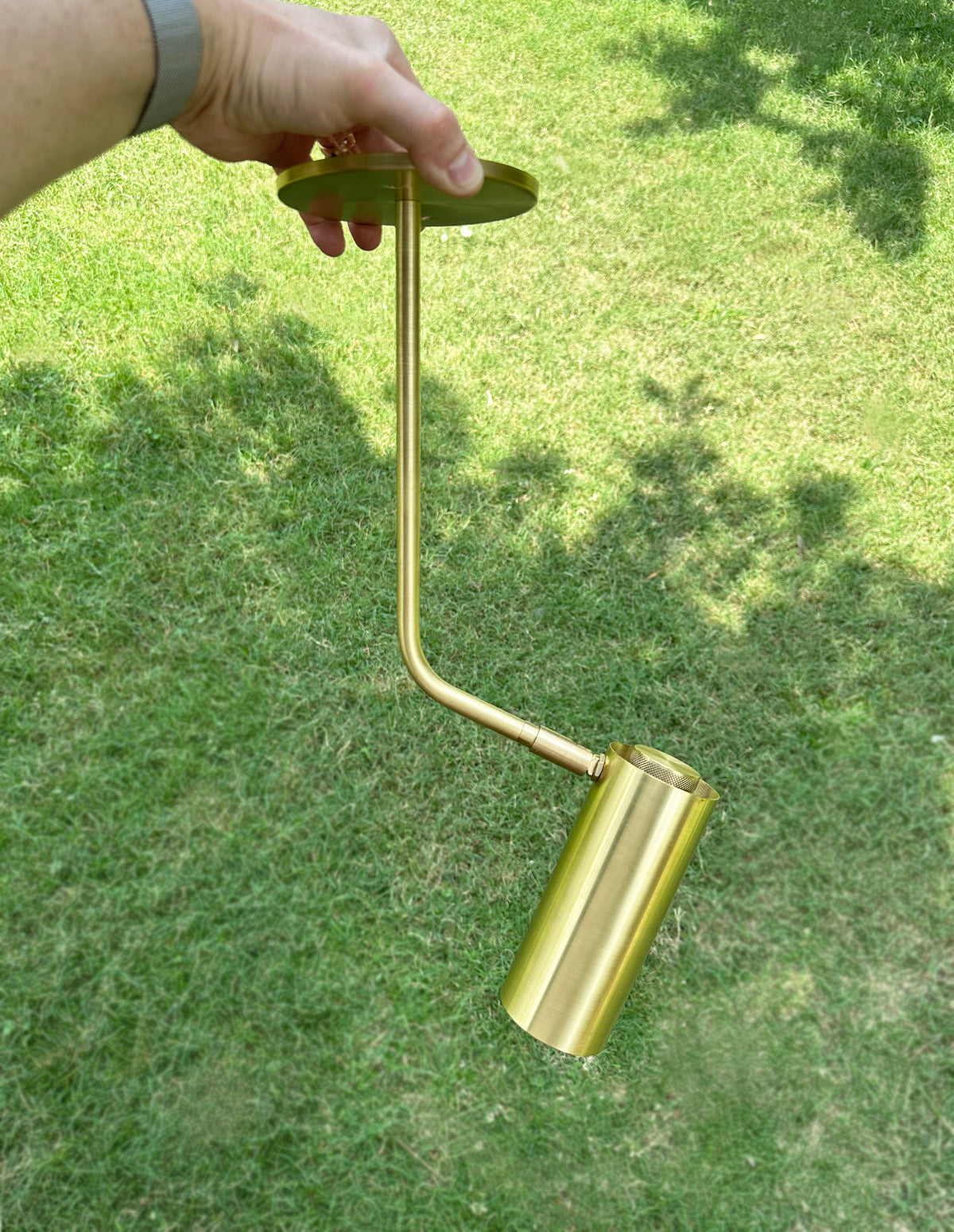 A hand holds the Brass Cylinder Bent Arm Drop Spot with Long Shade by Long Made Co., a brass-colored, cylindrical directional ceiling spotlight, outdoors on a grassy lawn in sunlight and shade.