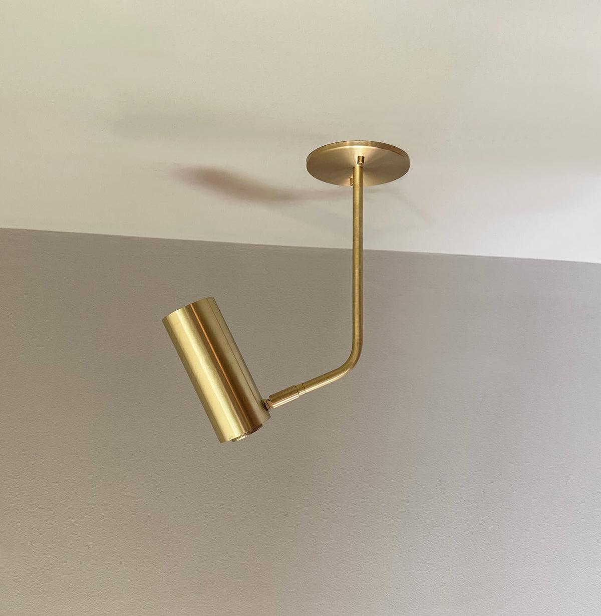The Brass Cylinder Bent Arm Drop Spot with Long Shade by Long Made Co. features a solid brass cylindrical shade and hangs from a round base, shown on a smooth, light-colored ceiling and wall background.