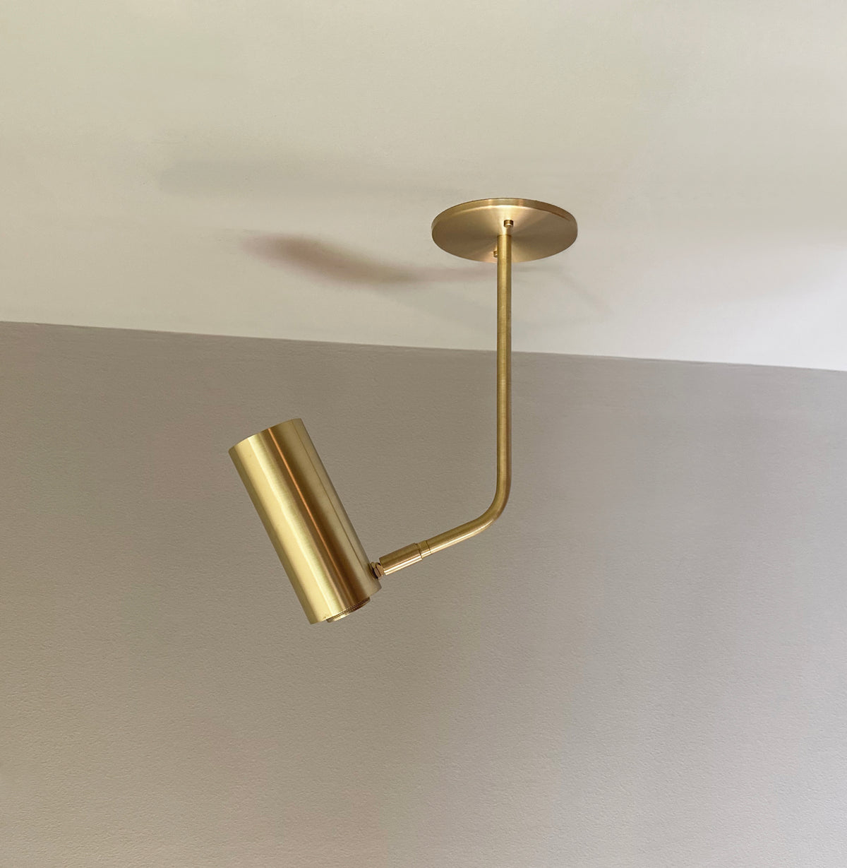 The Brass Cylinder Bent Arm Drop Spot with Long Shade by Long Made Co. features solid brass construction and a cylindrical shade, extending and angling outward. Set against a beige and taupe wall, it perfectly suits mid-century modern interiors.