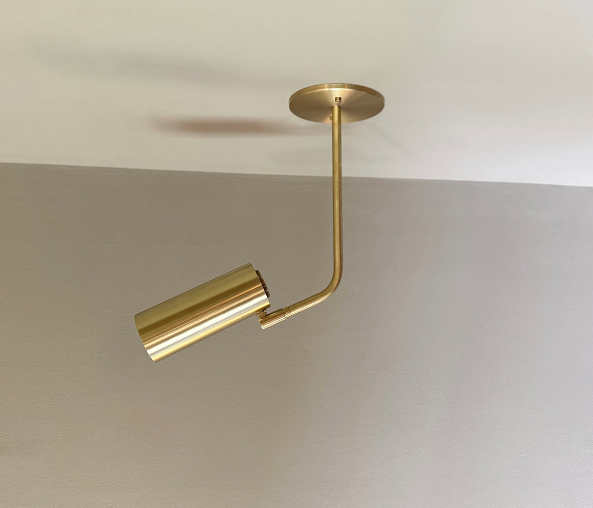The Brass Cylinder Bent Arm Drop Spot w/ Long Shade by Long Made Co. is a modern directional ceiling spotlight with a cylindrical shade and right-angled arm, crafted from solid brass, mounted on a white ceiling above a gray wall.