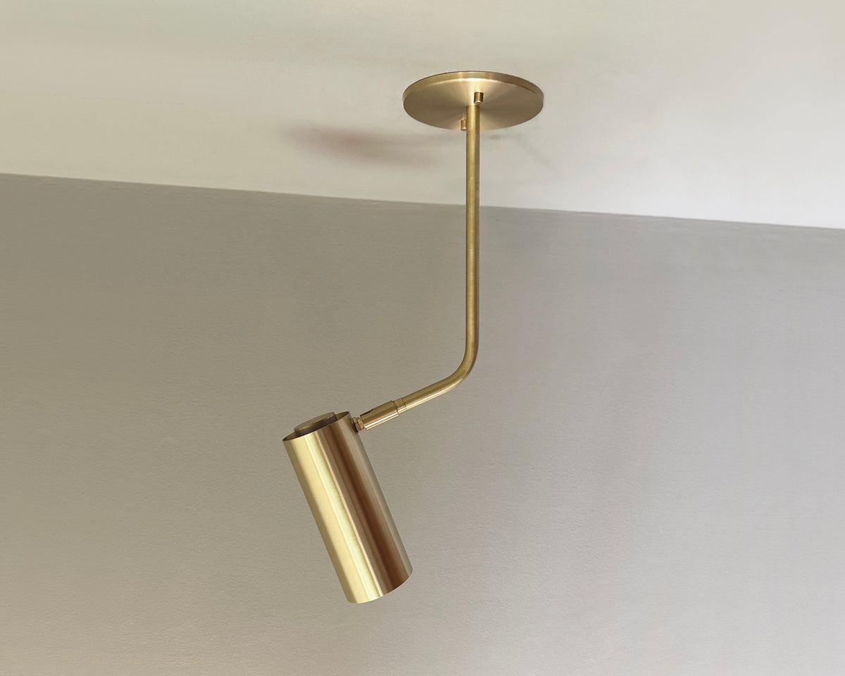 The Brass Cylinder Bent Arm Drop Spot with Long Shade by Long Made Co. is a gold-toned, solid brass directional ceiling spotlight with a downward angled arm, shown mounted on a light gray ceiling and wall.